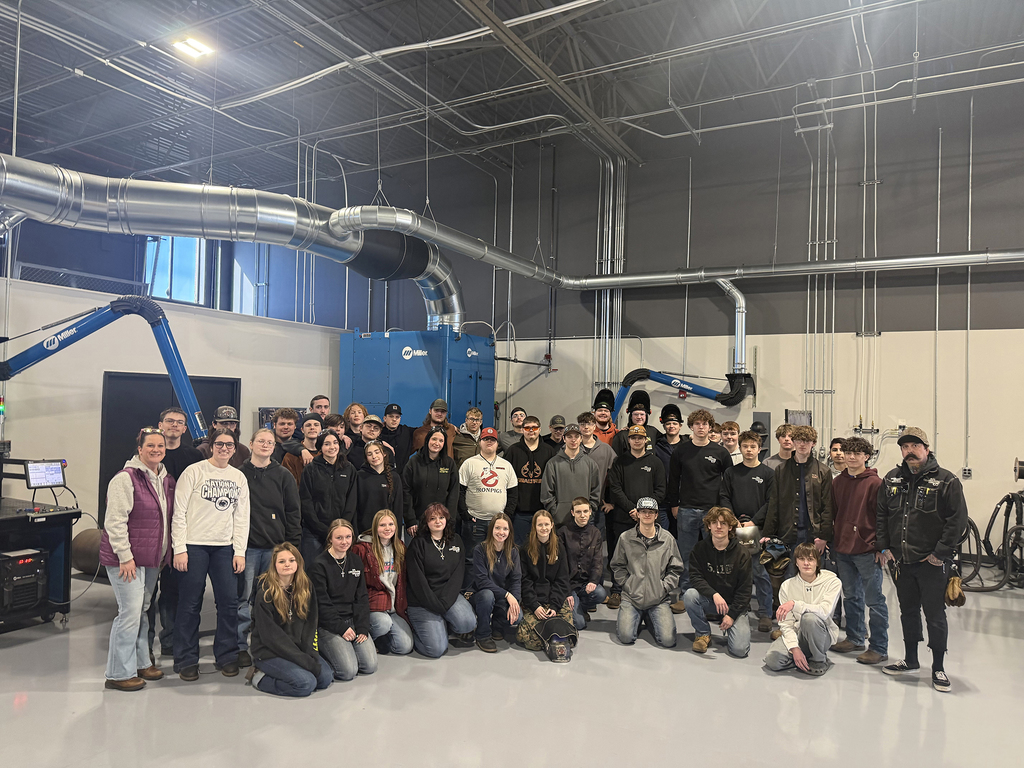Large group of welding students and instructors pose together inside a high-tech welding training facility with equipment and ventilation systems visible overhead.