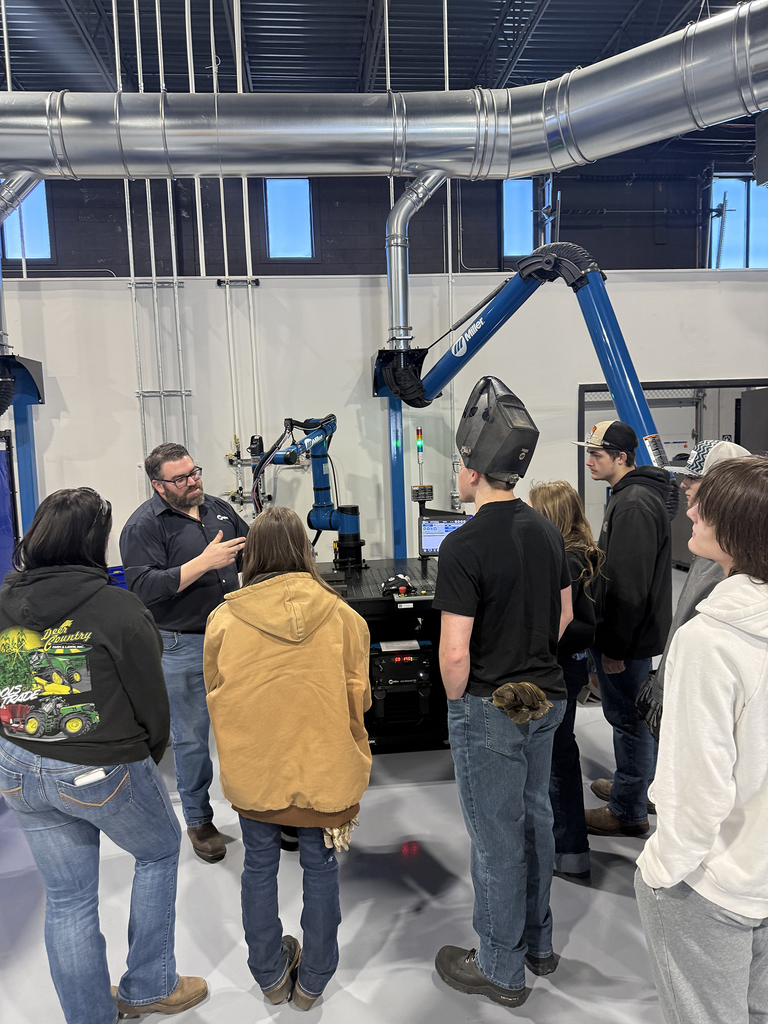 An industry instructor demonstrates robotic welding equipment to a group of students gathered around a workstation inside a welding training facility.