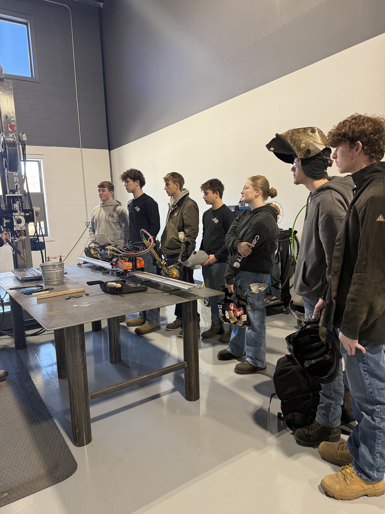 Welding students stand in a line beside a metal worktable, listening to instruction while holding welding helmets and gloves in a lab environment.