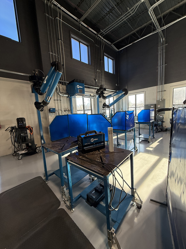 Wide view of a modern welding training lab with multiple blue welding booths, fume extraction arms overhead, and worktables set up with welding machines and metal pieces.