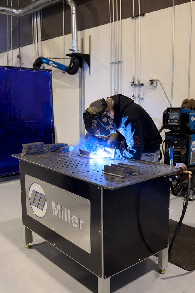 A student wearing a welding helmet and gloves welds a metal joint on a table, with sparks visible and metal practice pieces nearby.