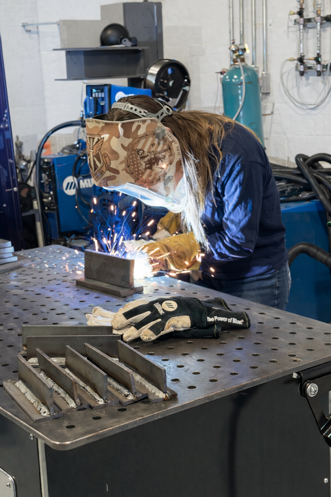 A student welds metal pieces on a perforated welding table while an instructor observes and gives guidance in a training lab.