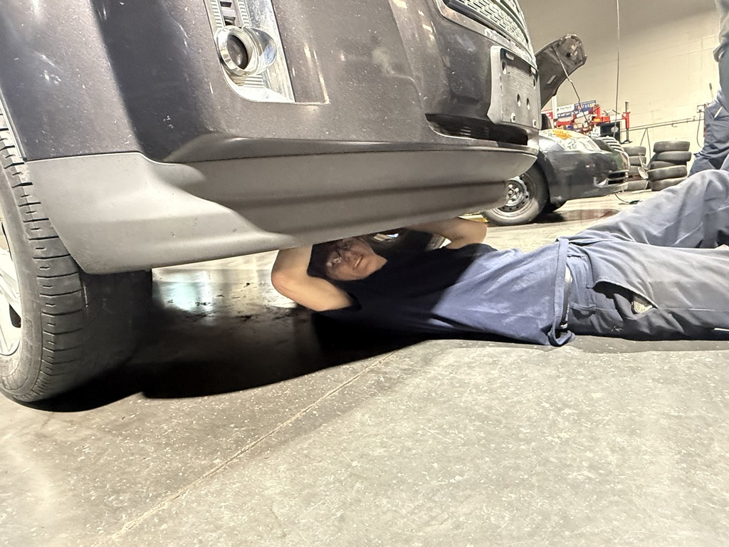 Student lies on the shop floor beneath the front of a vehicle, working on components underneath the car as part of hands-on automotive training.