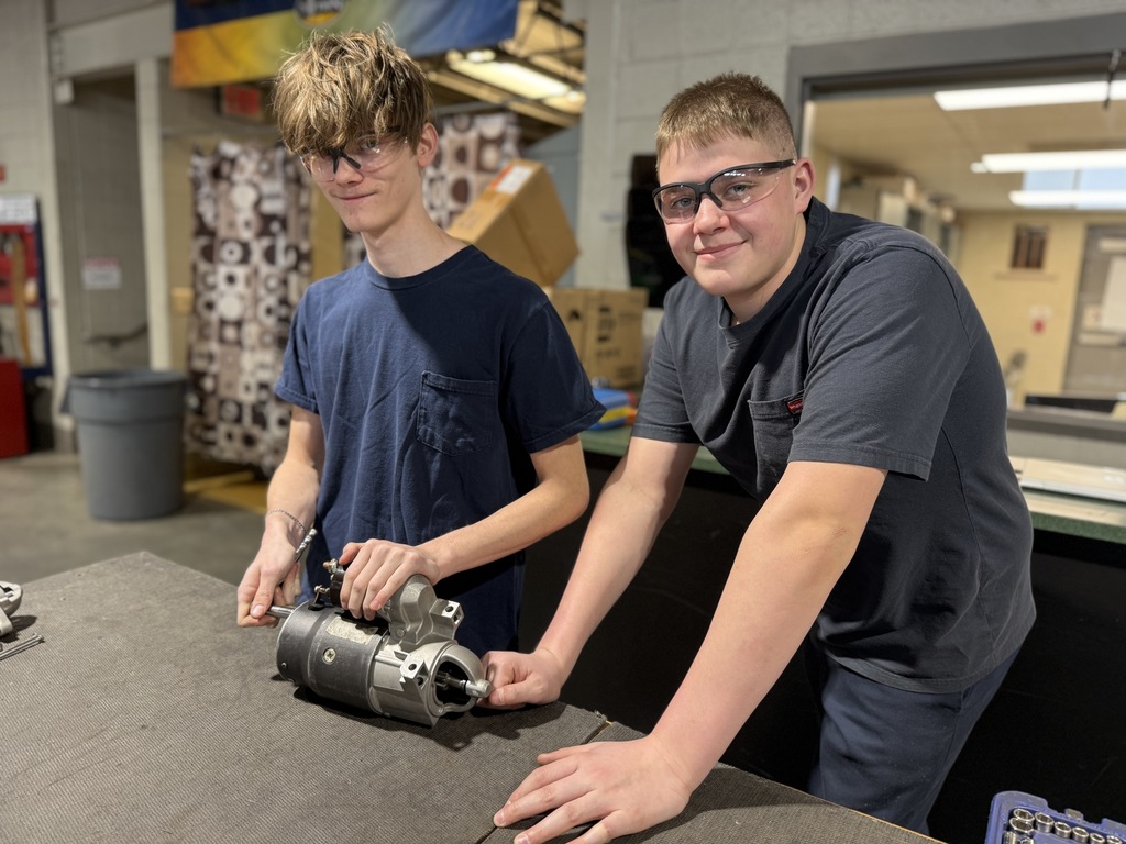 Two Automotive Technology students wearing safety glasses stand at a workbench with a starter motor, tools, and equipment while preparing or inspecting parts.