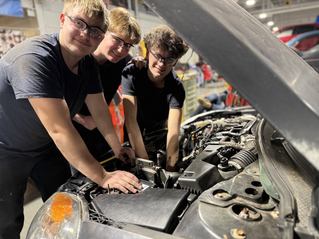 Three Automotive Technology students lean over an open engine bay, working together on components under the hood during a hands-on automotive repair activity.