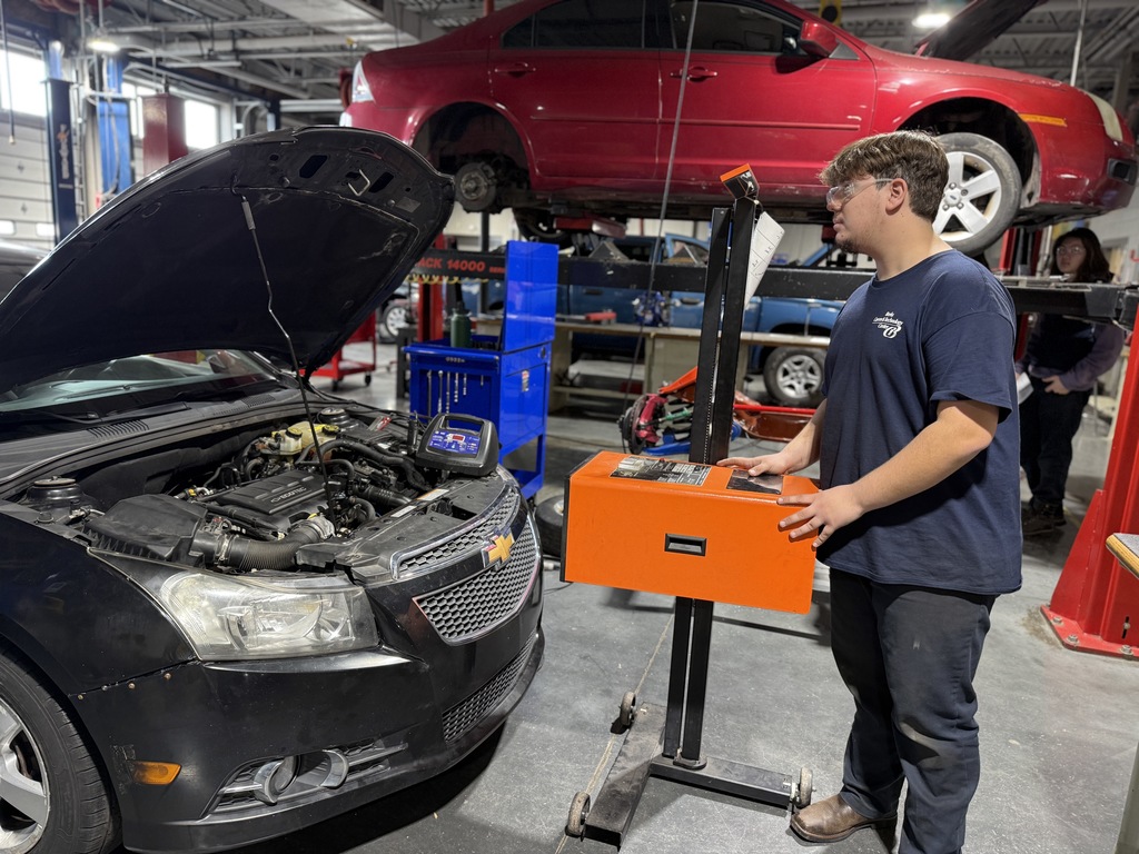 Student in safety glasses uses a diagnostic cart while checking the engine of a black car with the hood open in an automotive shop; another vehicle is lifted on a hoist in the background.