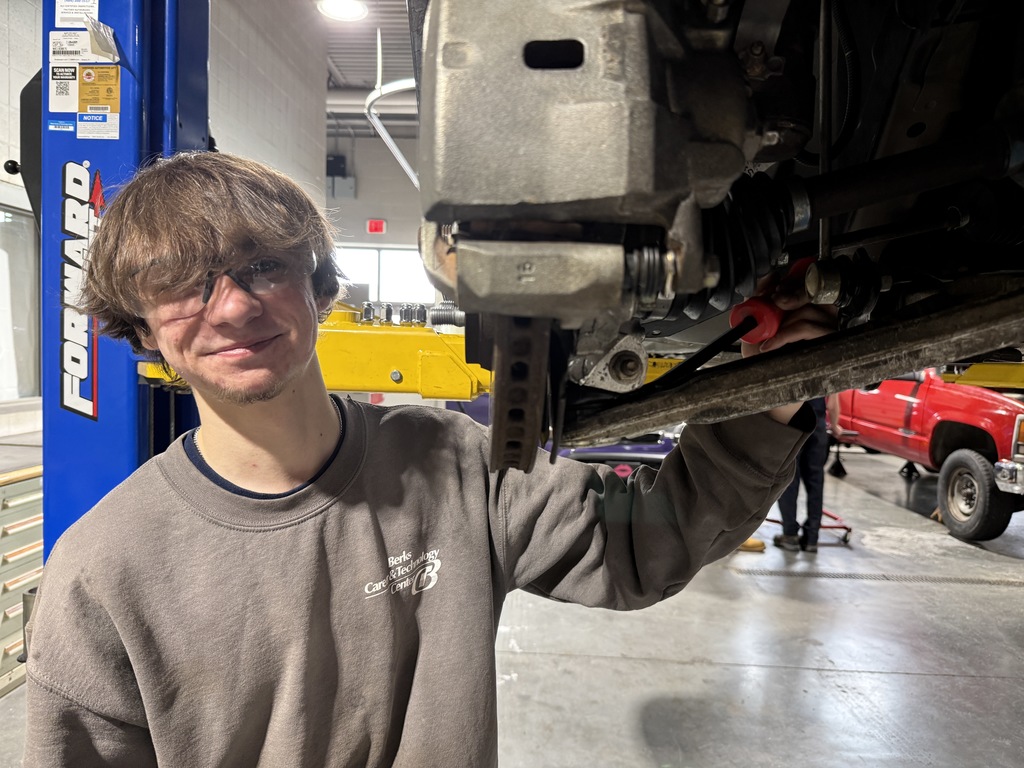 Automotive student wearing safety glasses works underneath a raised vehicle, using a tool near the suspension and brake assembly inside a shop with vehicle lifts.