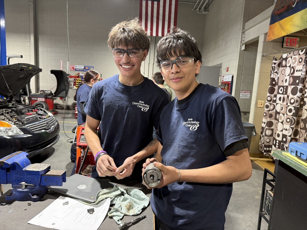 Two Automotive Technology students wearing safety glasses stand at a workbench, smiling while holding a vehicle starter component and small parts during a repair activity.