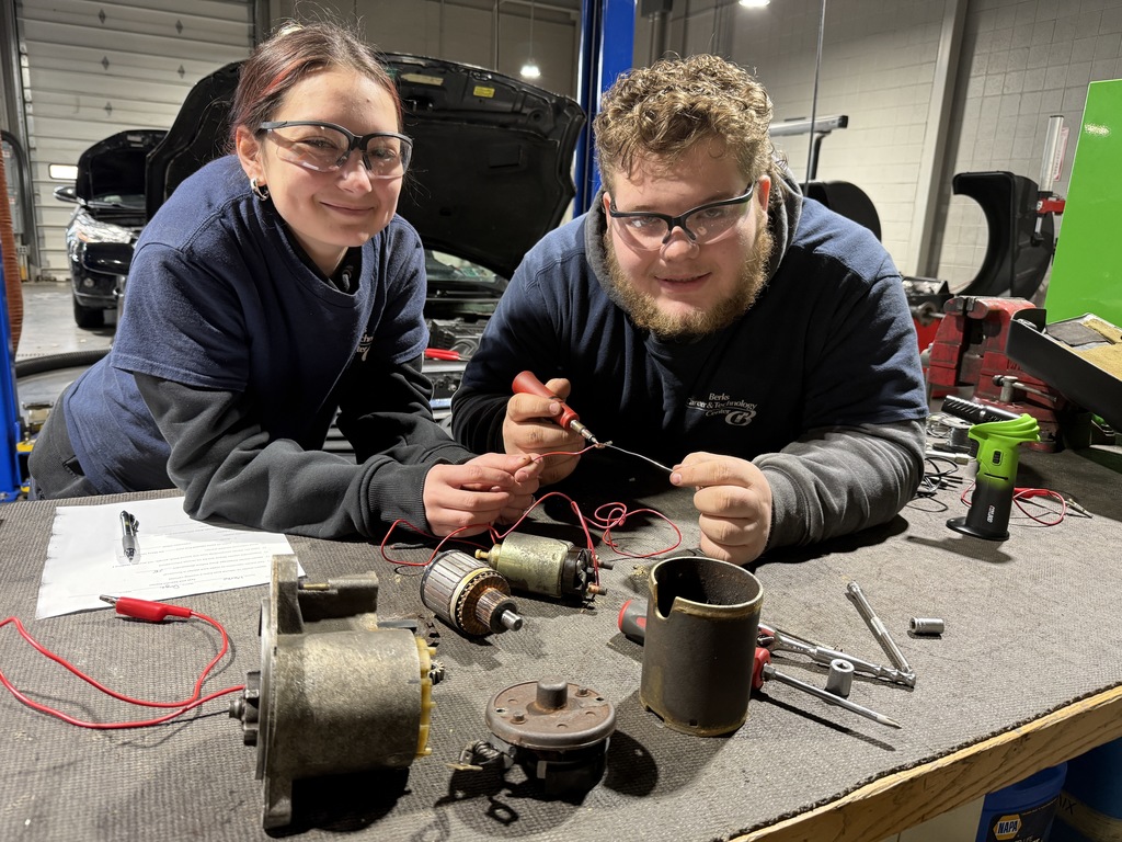 Two Automotive Technology students wearing safety glasses sit at a workbench with disassembled starter motor parts, using a test lead and tools during an electrical repair lesson.