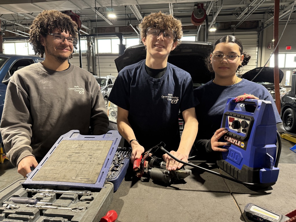 Three Automotive Technology students wearing safety glasses stand behind a workbench displaying tools, a starter motor, and a portable battery charger inside an auto shop.