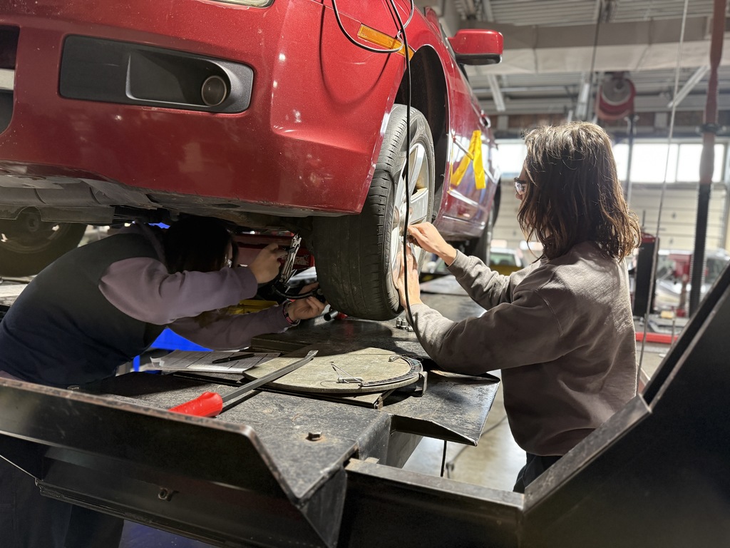 Two students work on a vehicle raised on a lift, using tools near the wheel area while performing service or inspection tasks in the automotive lab.