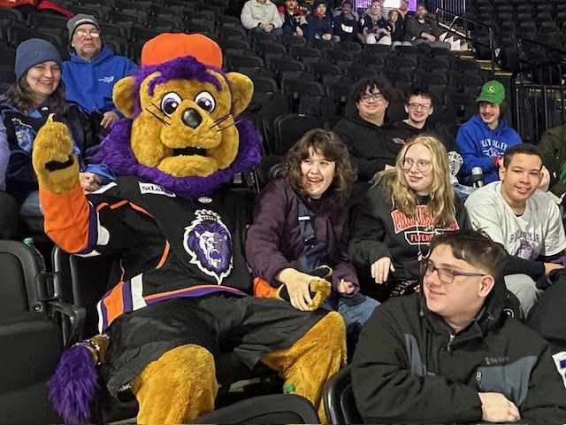 A group of students sit in arena seats next to the Reading Royals mascot, who is dressed in a purple and black hockey uniform and posing with them during the game.