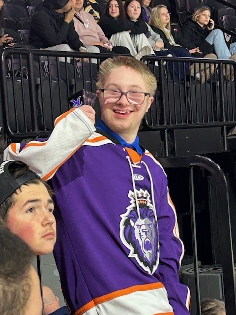 A student wearing a purple Reading Royals hockey jersey smiles and raises a foam finger while seated in the stands at an indoor hockey arena, with other spectators visible behind him.