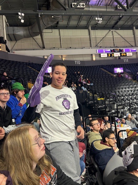 A student standing in the arena seating area holds up a purple foam finger and smiles, wearing a light-colored Reading Royals shirt, while other fans sit nearby watching the game.