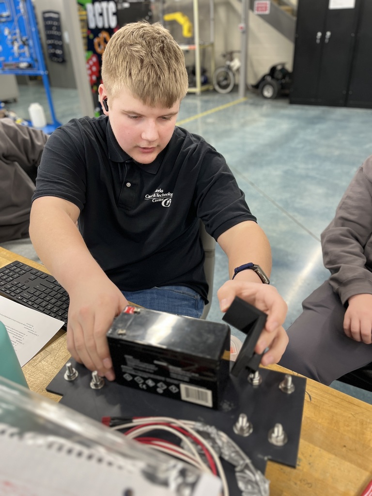 A Mechatronics Engineering Technology student installs a rectangular battery onto a mounted base plate, aligning it with bolts and wiring as part of an electrical system assembly at a classroom workbench.