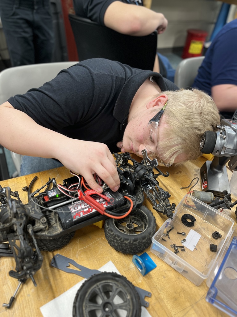 A Mechatronics Engineering Technology student wearing safety glasses leans over a remote-controlled vehicle chassis, adjusting internal wiring and components while small tools and parts are spread across the workbench.