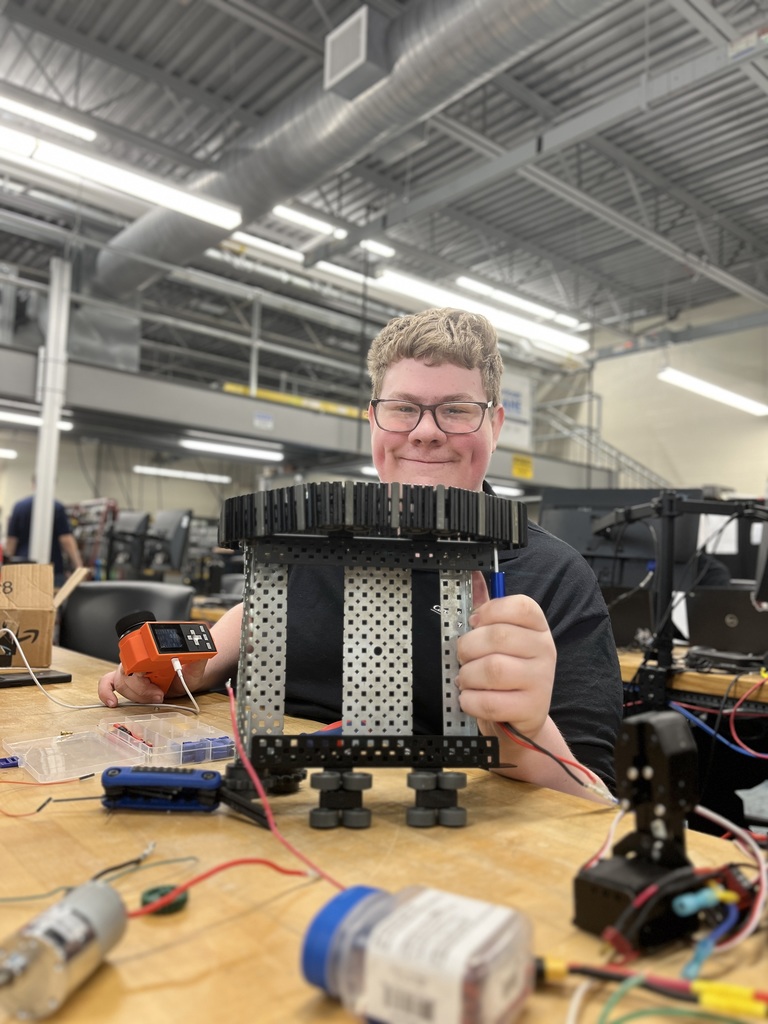 A Mechatronics Engineering Technology student sits at a workbench holding a partially assembled mechanical structure with gears and metal supports, surrounded by wiring, tools, and electronic components in a lab classroom.