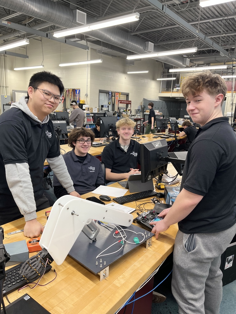 Four Mechatronics Engineering Technology students stand and sit around a workbench in a lab, posing with a robotic arm assembly mounted on a base, with computer monitors, wiring, and tools visible on the table.