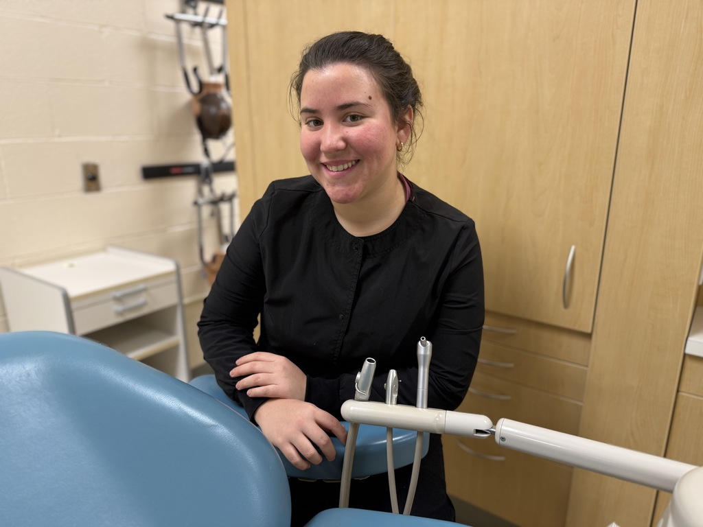 Lauren Oliva, a Dental Occupations student, smiles while standing beside a dental chair in a clinical lab, with dental instruments visible in the foreground and cabinetry in the background.
