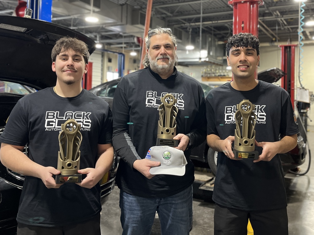 Two Automotive Technology students and their instructor stand in an automotive lab, holding third-place trophies from a regional technician competition. The group is wearing matching black program shirts, with vehicle lifts and cars visible in the background.