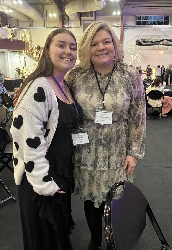 A Berks Career & Technology Center student stands smiling next to a Night to Shine guest, both wearing name badges inside an event venue decorated with lights.
