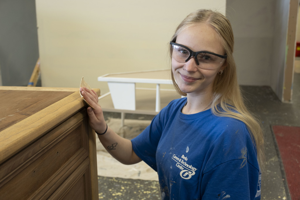 Student wearing safety glasses sands the edge of a wooden cabinet in a Berks Career & Technology Center workshop, focusing on surface finishing.