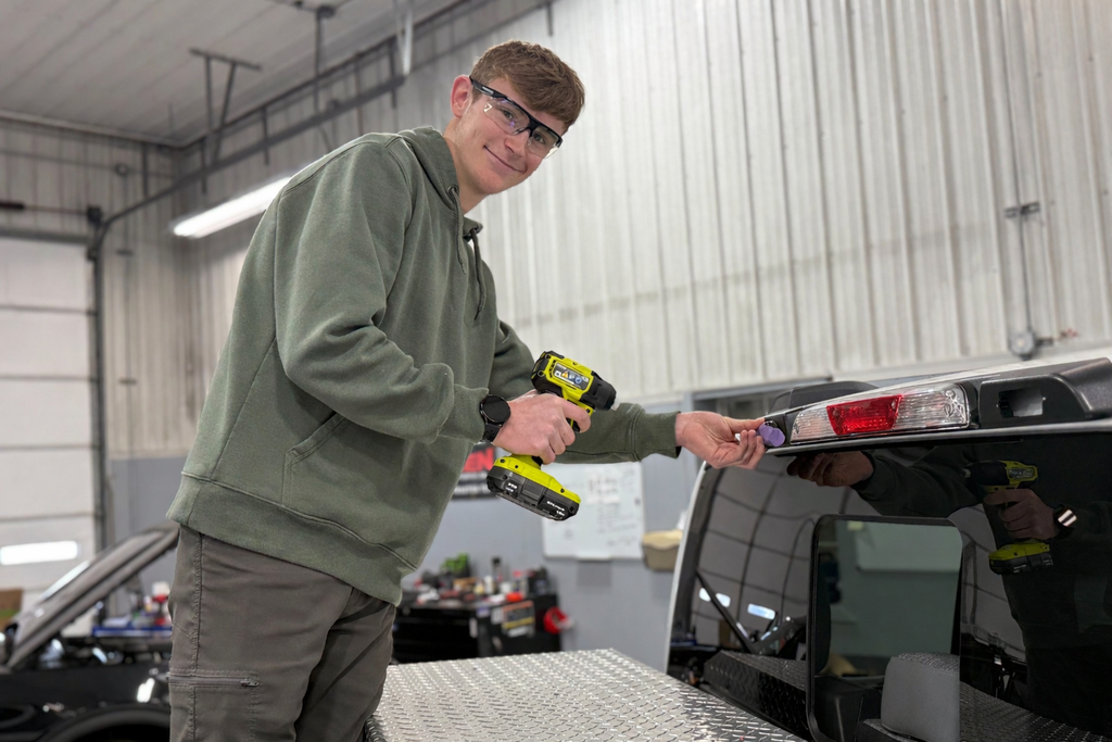 Student wearing safety glasses uses a cordless drill to work on the rear light area of a pickup truck inside an automotive shop, standing beside the truck bed with tools and equipment visible in the background.