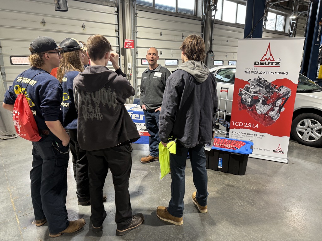 A group of BCTC students listens as an industry professional explains career opportunities at a transportation-focused exhibit featuring diesel engine components and tools.