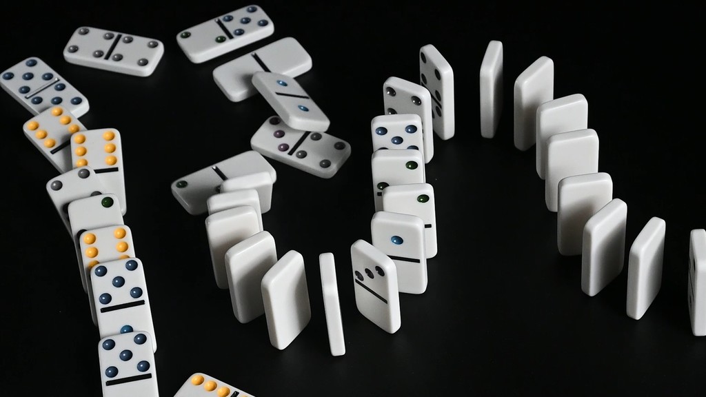 White domino tiles arranged in multiple curved lines on a dark background, with some dominoes standing upright and others fallen, visually representing a chain reaction.