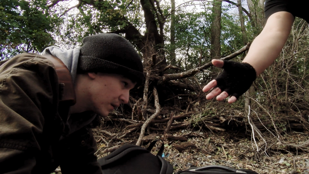 A young person sitting on the ground in a wooded area looks up as another person extends a hand toward them, symbolizing help, support, and connection.