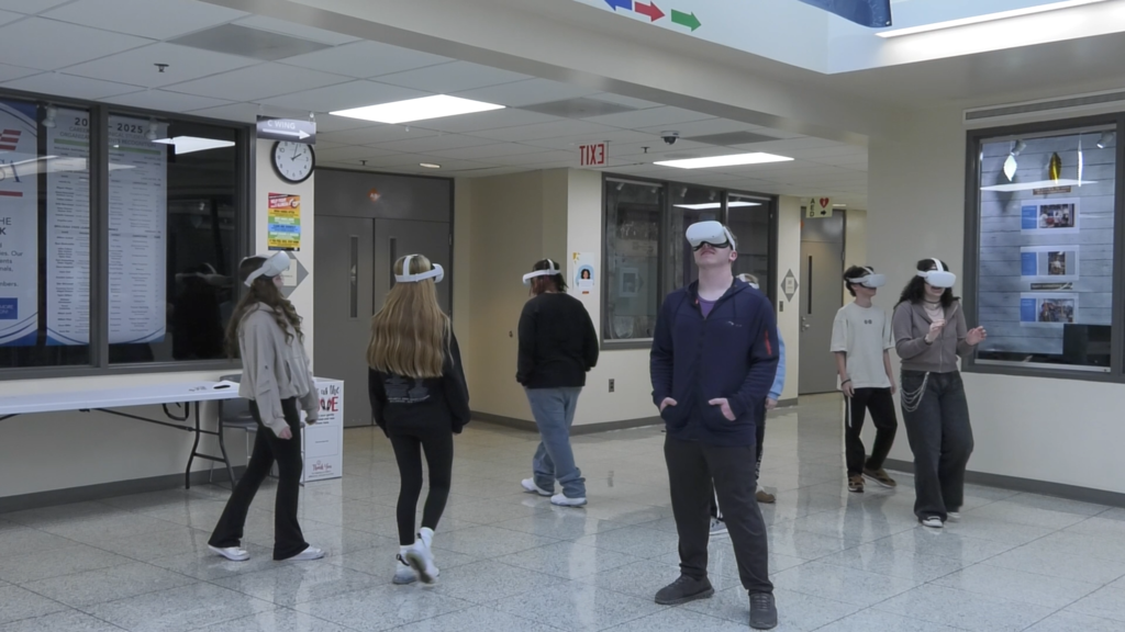 Students wearing virtual reality headsets stand and walk through a school hallway, participating in an immersive activity that explores perspective and experience.