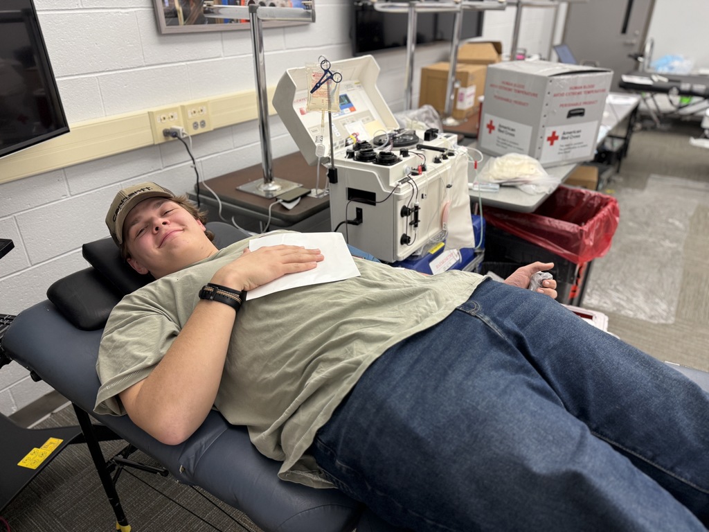 A student lies on a donation bed during a blood drive, smiling while connected to blood collection equipment in a classroom-style setting.