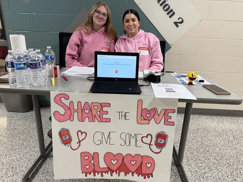 Two students sit behind a check-in table at a blood drive with a laptop, water bottles, and a handmade “Share the Love, Give Some Blood” sign displayed in front.