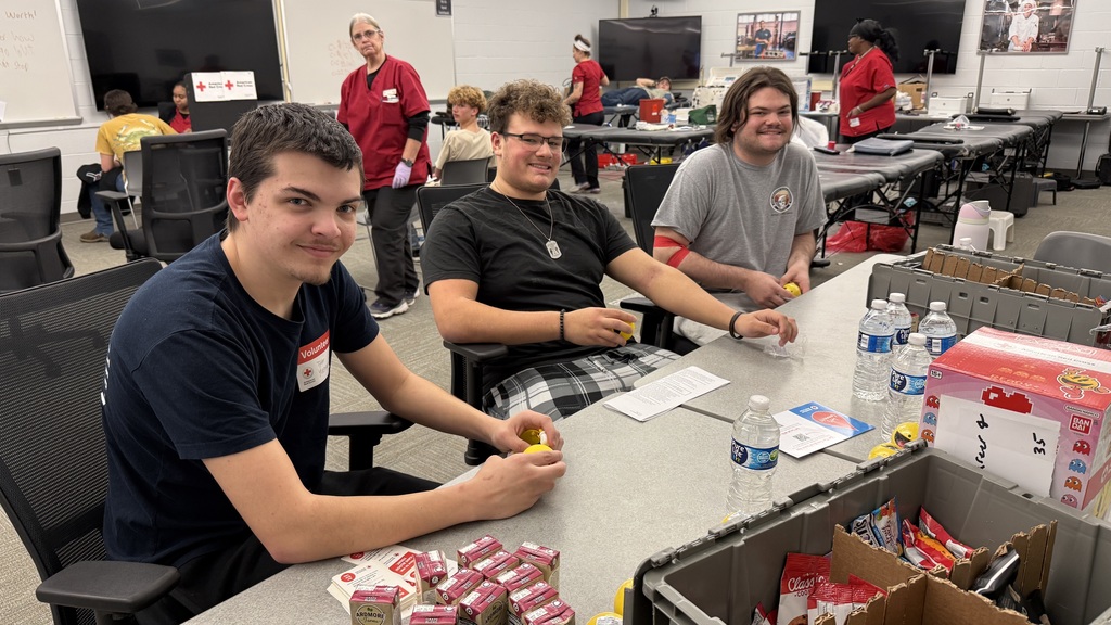 Three students sit at a table holding snacks and drinks after donating blood, with medical staff and donation stations visible in the background.