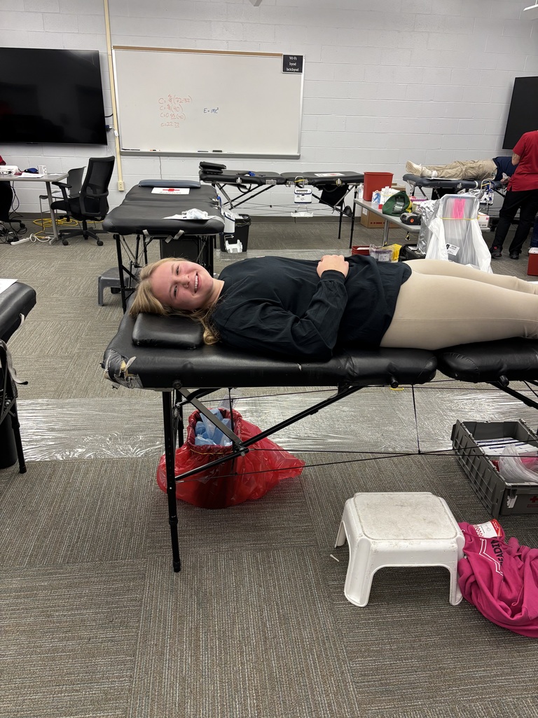 A student lies on a donation bed and smiles toward the camera during a blood drive, with medical equipment and other donation stations visible behind her.