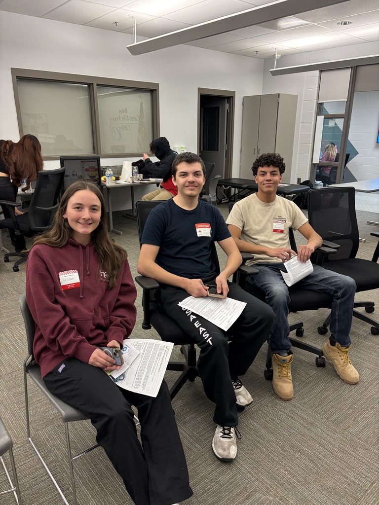 Three students sit in chairs holding paperwork and personal items while waiting during a blood drive, with tables and other participants visible in the background.