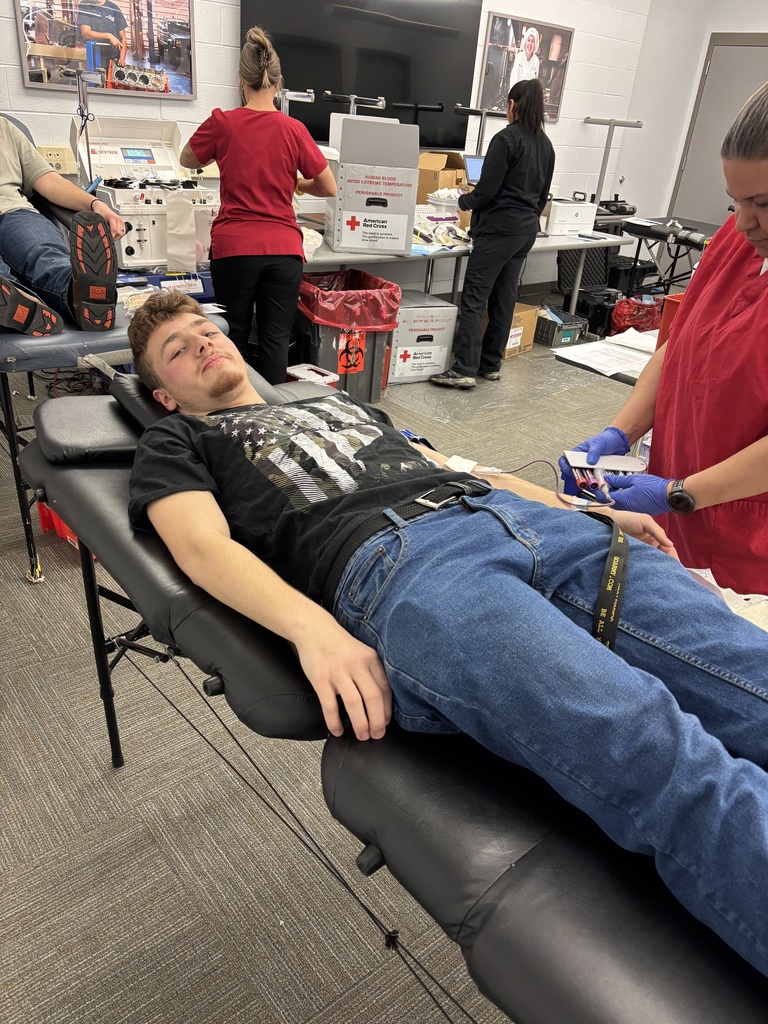 A student lies on a donation bed while a Red Cross staff member prepares blood collection equipment, with additional donation stations and staff visible in the room.