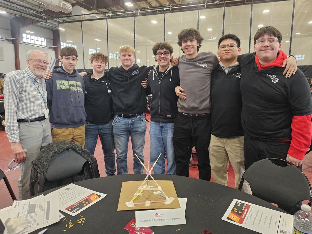 Mechatronics Engineering Technology students stand together smiling behind a table with a completed engineering structure made from lightweight materials during a team challenge.