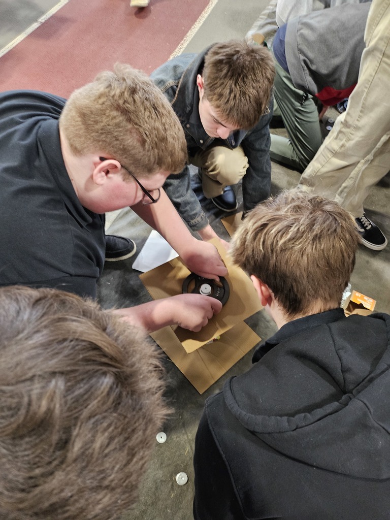 Several Mechatronics Engineering Technology students kneel closely together while testing the strength of a small tower structure by placing weighted plates on top.