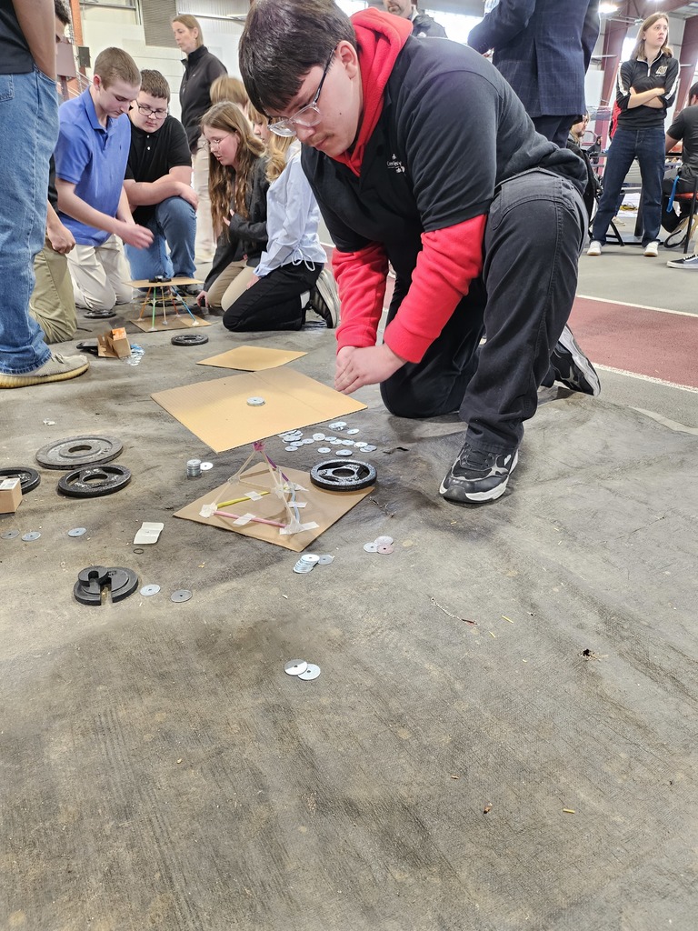 A Mechatronics Engineering Technology student kneels on the floor carefully adding metal weights to a small tower structure as classmates observe during a hands-on engineering challenge.