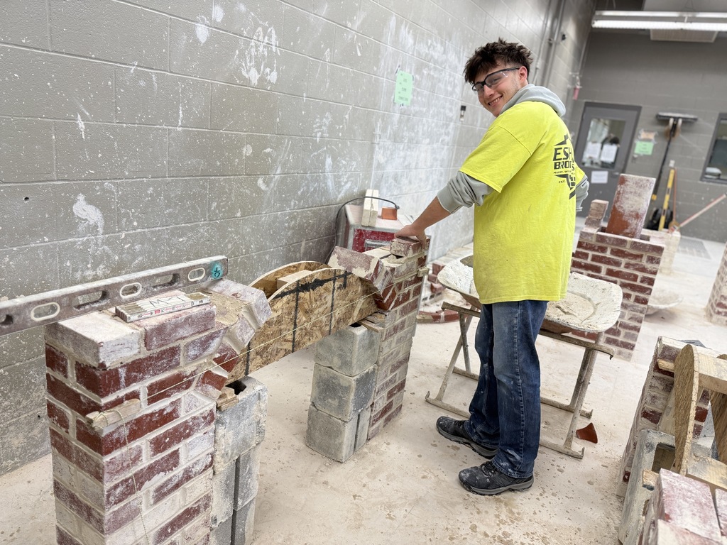 Masonry student sets bricks on a small arch form, smiling toward the camera in the lab.