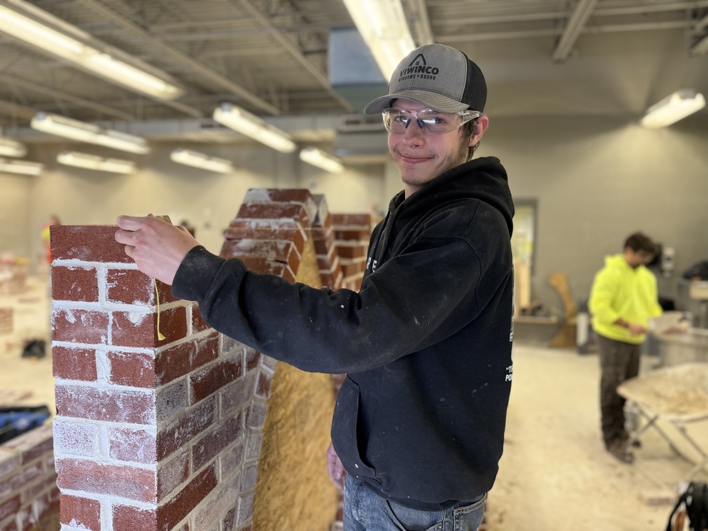 Masonry student wearing safety glasses measures the top of a brick column inside a construction lab.