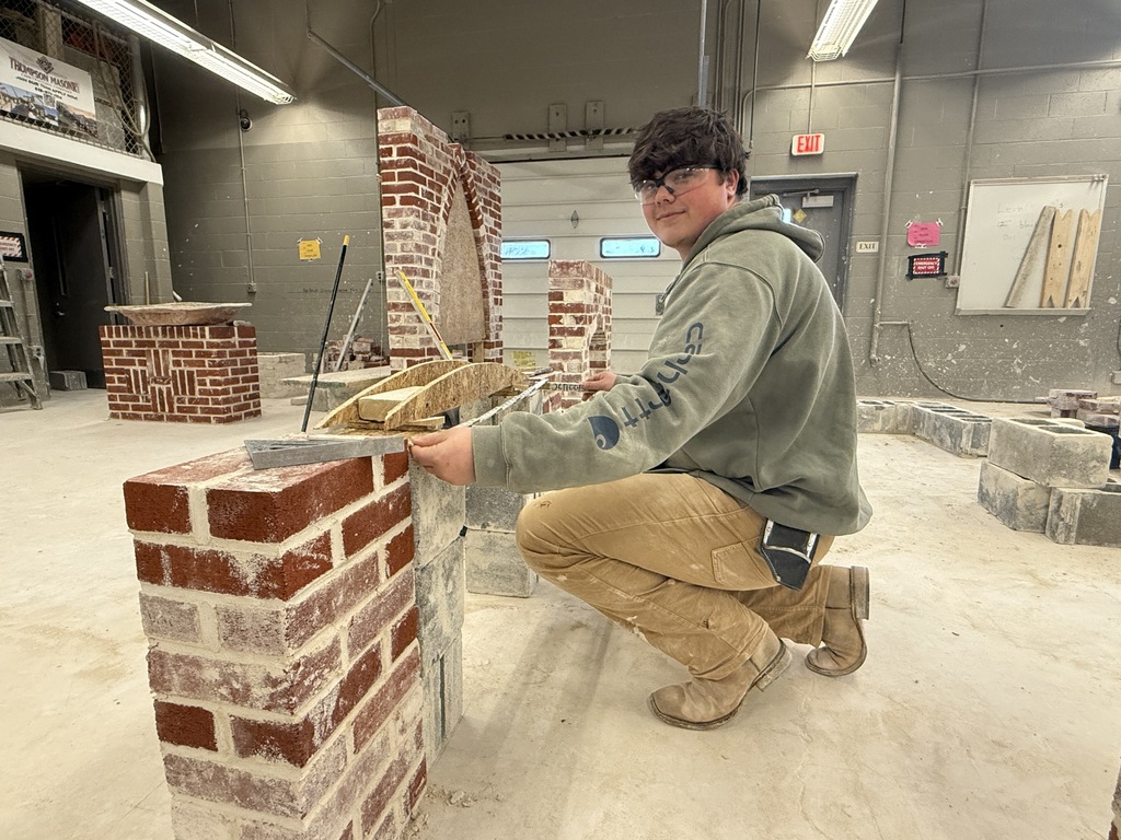 Masonry student kneels beside a brick arch form, checking alignment with a hand tool.