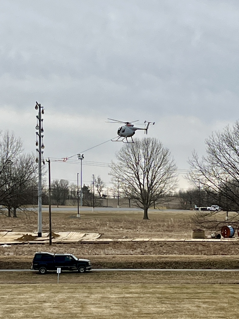 Helicopter flies low while guiding transmission lines across an open field near utility poles.