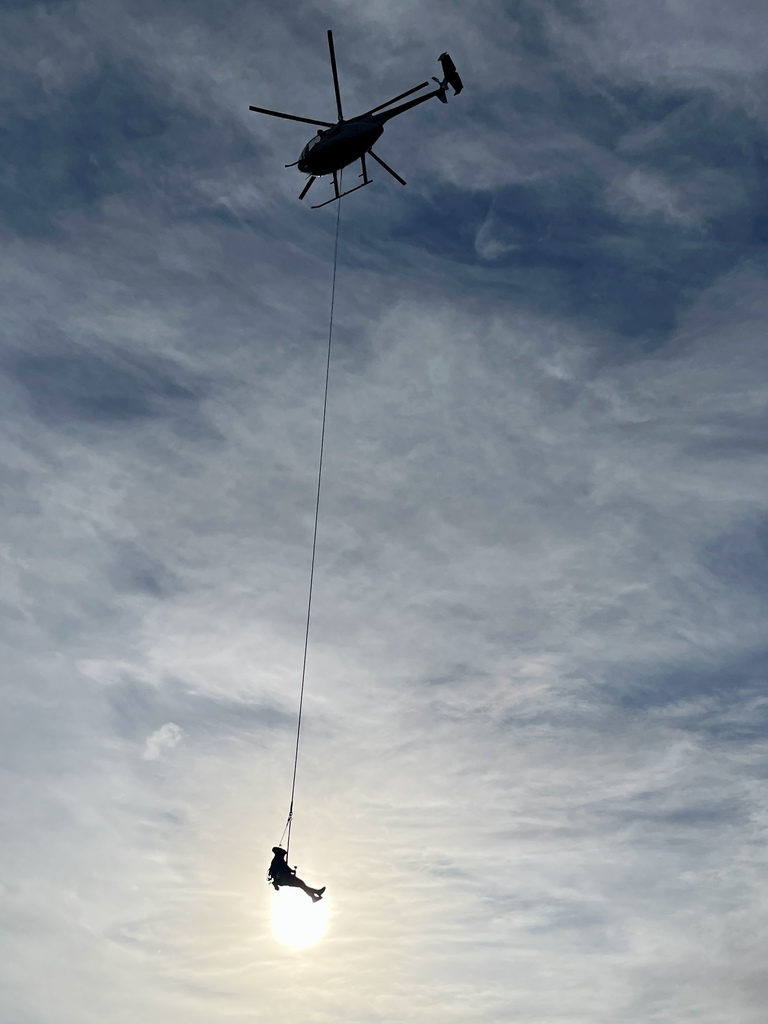 Helicopter installing transmission lines using a suspended guide rope against a cloudy sky.