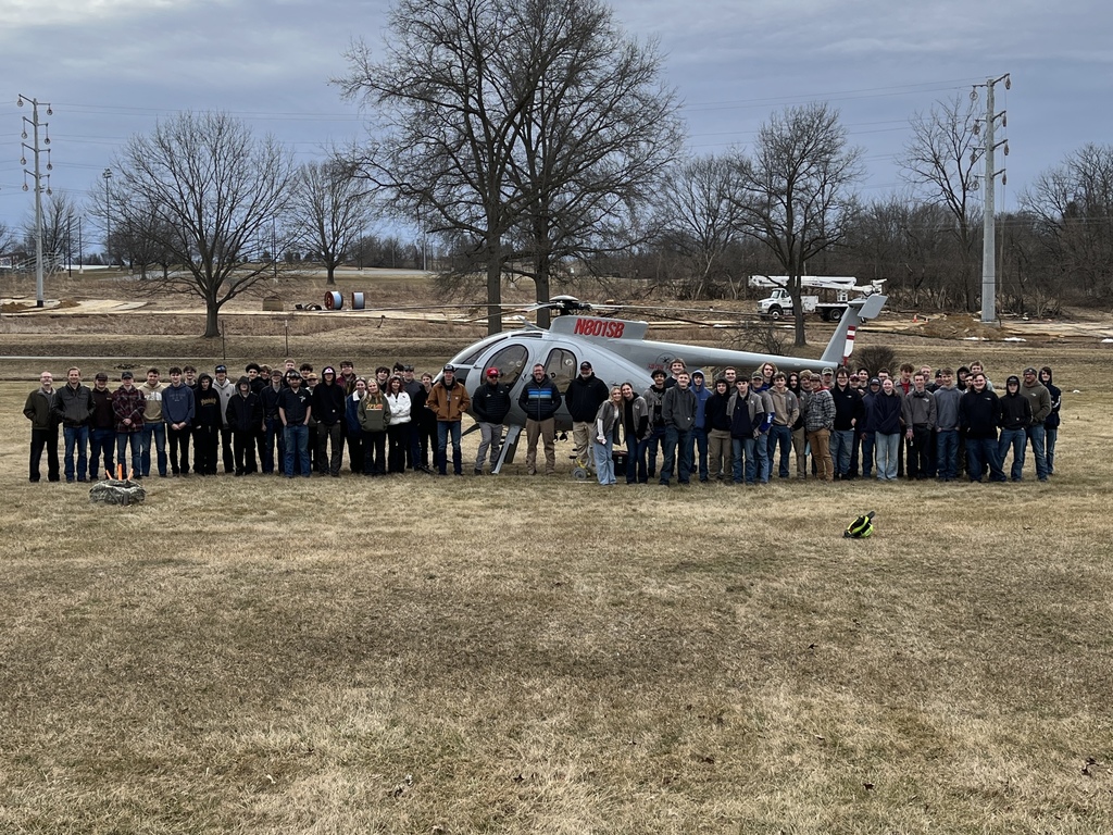 Wide group photo of students and industry partners standing in front of a helicopter at the project site.