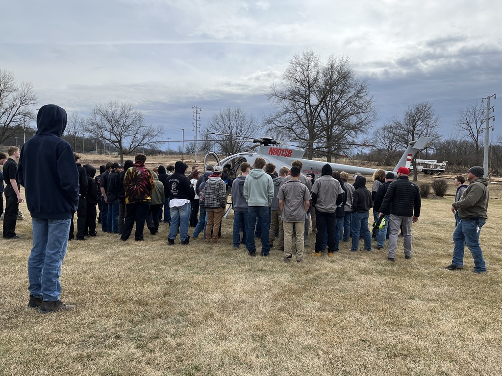 Students gather around a helicopter on the ground as professionals explain the equipment and process.