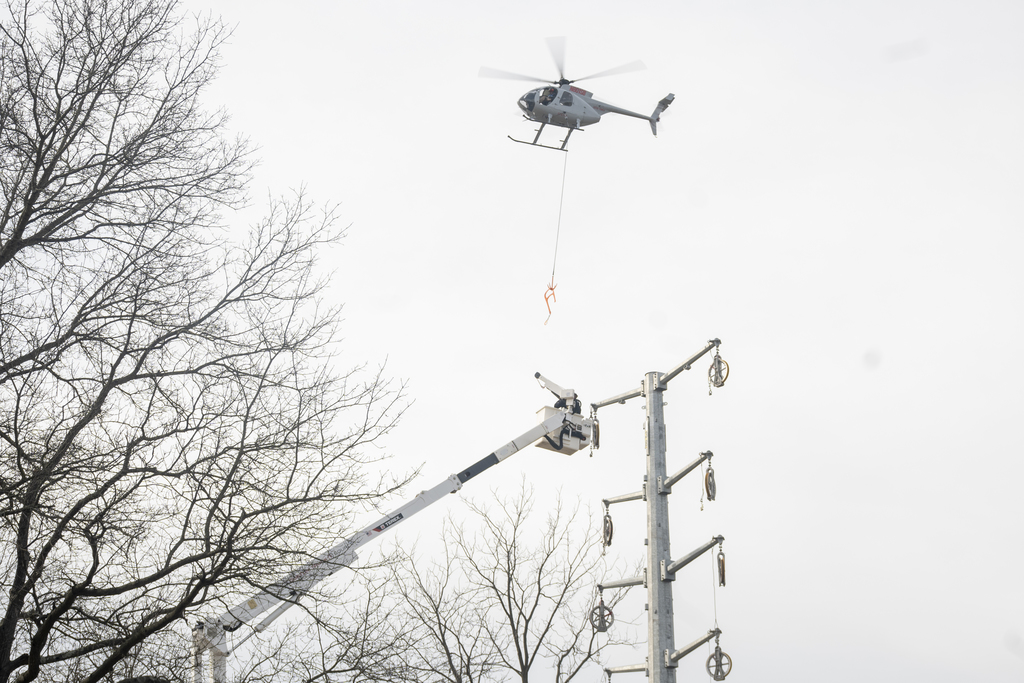 Helicopter hovers above a field while a worker manages a guide rope below during line installation.