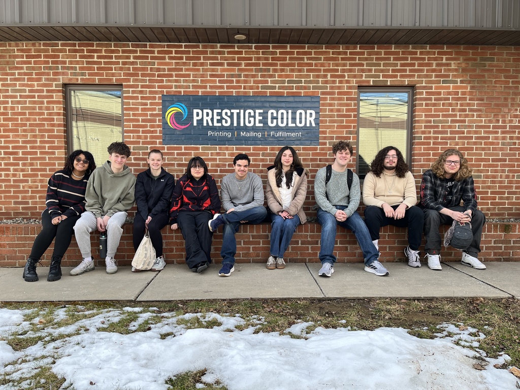 Advertising Art & Design Technology students sit along a brick ledge outside Prestige Color printing facility in Lancaster beneath a sign that reads “Prestige Color – Printing | Mailing | Fulfillment.”