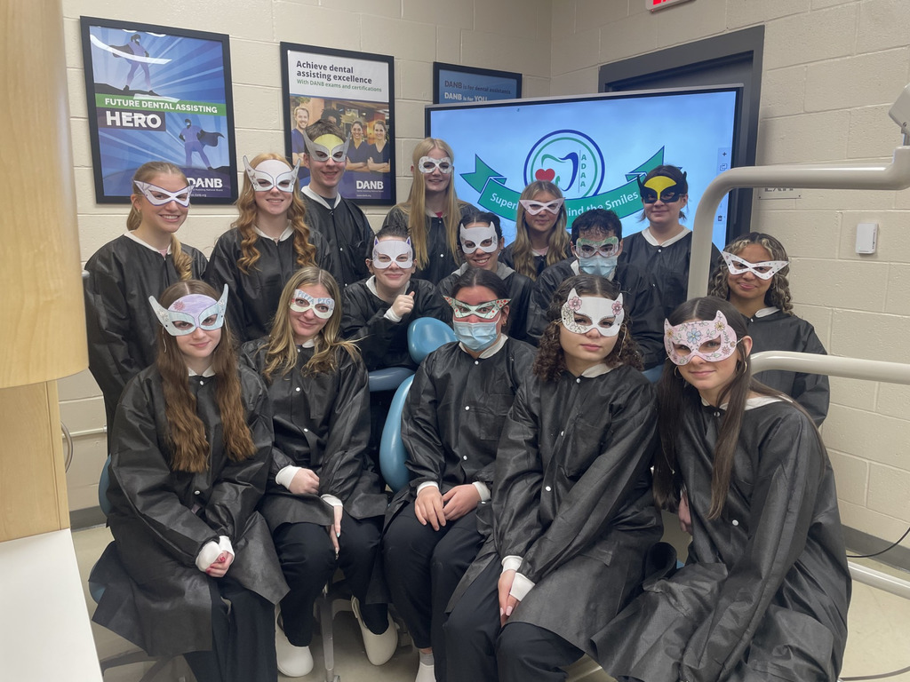 Dental Occupations students wearing superhero-style masks pose together in a dental lab classroom beside a screen displaying the “Superheroes behind the Smiles” Dental Assistants Recognition Week graphic.